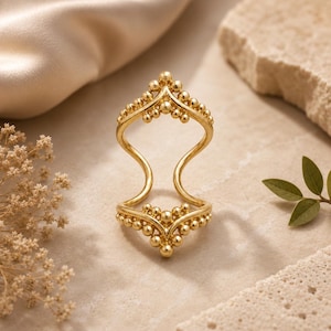 May include: A gold-toned metal ring featuring an open, elongated design that spans two finger joints. The ring is composed of two V-shaped bands adorned with small, spherical bead accents along the edges, connected by two smooth, curved side bars.