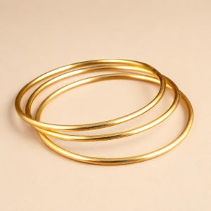 Può includere: Un set di tre bracciali rigidi sottili e circolari in metallo color oro. Ogni bracciale presenta una superficie liscia e lucida con un profilo rotondo costante. I tre bracciali sono impilati e leggermente sovrapposti tra loro.