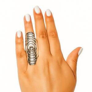 Puede incluir: Un anillo plateado con un diseño único de círculos concéntricos. El anillo se lleva en una mano con esmalte de uñas blanco. El diseño del anillo es alargado y de forma ovalada, con un círculo central elevado.