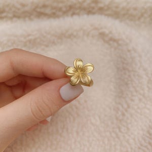Op de afbeelding: Een goudkleurige ring met een gedetailleerd bloemmotief. De bloemblaadjes van de ring zijn getextureerd en de band is glad. De ring wordt tegen een zachte, neutrale achtergrond gehouden.