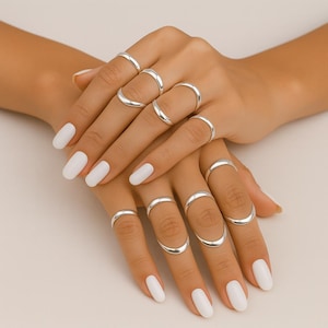 Può includere: Una collezione di anelli d'argento in vari stili adorna un paio di mani con smalto bianco. Gli anelli includono semplici fasce e design curvi, mostrando un'estetica di gioielli minimalista. Gli anelli sono esposti su uno sfondo chiaro.