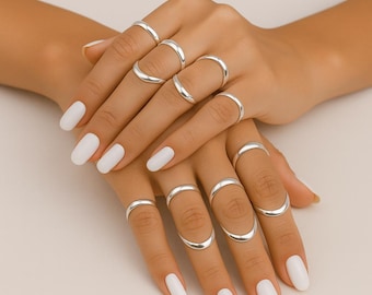 May include: A collection of silver rings in various styles adorns a pair of hands with white nail polish. The rings include simple bands and curved designs, showcasing a minimalist jewelry aesthetic. The rings are displayed on a light background.