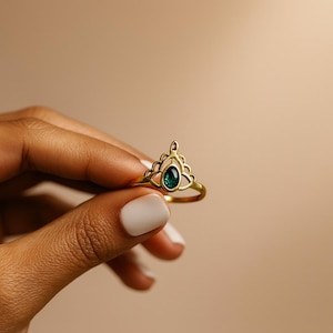 Pode incluir: Um anel dourado com uma pedra preciosa verde esmeralda. O anel apresenta um design decorativo vazado em forma de coroa. A pedra preciosa oval está no centro do anel.