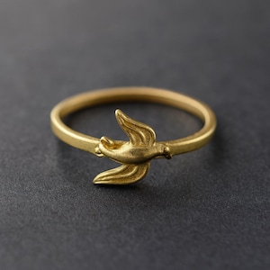 Op de afbeelding: Een goudkleurige ring met een gedetailleerd vogelontwerp. De vogel is in vlucht afgebeeld, met gespreide vleugels. De ringband is een eenvoudige, gladde cirkel. De ring staat tegen een donkergrijze achtergrond.
