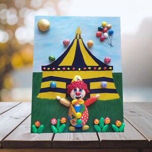 May include: A colorful, handcrafted circus-themed artwork featuring a clown in front of a yellow and black striped tent. The artwork includes balloons, balls, and flowers, all set against a blue sky and green grass background.
