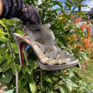 May include: Clear, open-toe high-heeled sandals with silver platforms and stiletto heels. The sandals feature a cage-like design adorned with small, sparkling embellishments. The heels have a red sole and are approximately 10 cm tall.
