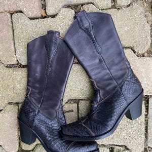 Peut inclure: Paire de bottes de cowboy en cuir noir. La partie inférieure des bottes présente un motif peau de serpent, tandis que la partie supérieure est en cuir lisse. Elles ont un bout pointu, un talon bloc et des coutures décoratives.
