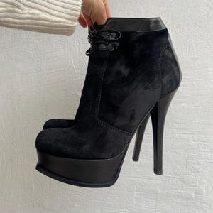 May include: Black suede platform ankle boots with high stiletto heels. The boots have a rounded toe and a leather trim at the top. A small decorative tie detail is visible near the ankle.