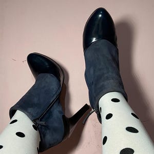 May include: A pair of navy blue ankle boots with a pointed toe and a high heel. The boots have a suede upper and a leather toe cap. The boots are paired with white tights with black polka dots.