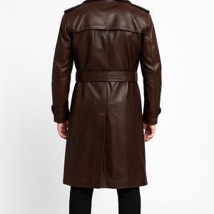 May include: A brown leather trench coat with a belted waist and a classic collar. The coat has long sleeves and a button closure. The model is wearing black pants and shoes. The coat is approximately 55 inches long.