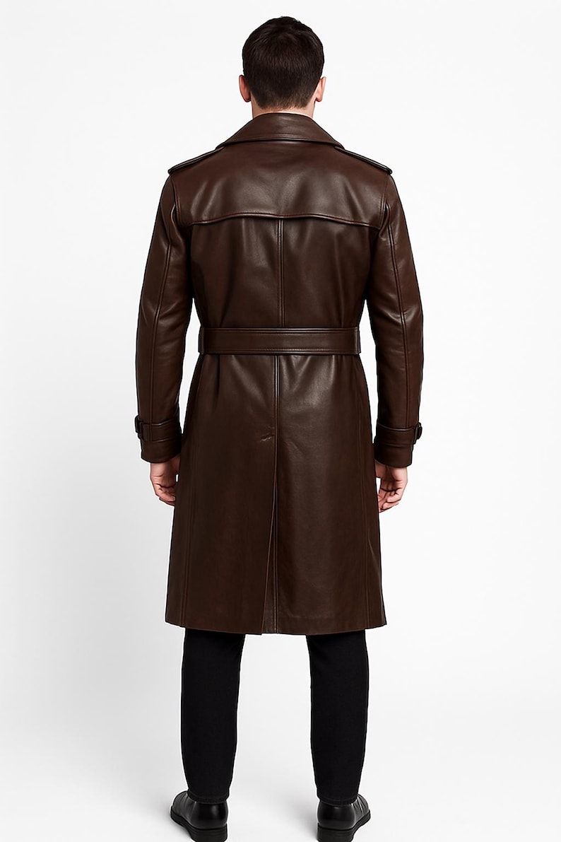 May include: A brown leather trench coat with a belted waist and a classic collar. The coat has long sleeves and a button closure. The model is wearing black pants and shoes. The coat is approximately 55 inches long.