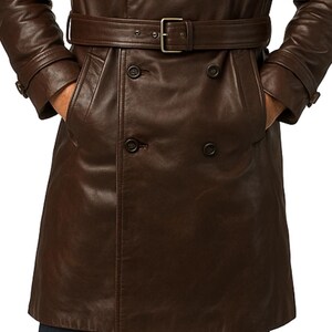May include: A brown leather trench coat with a double-breasted design. The coat features a belt with a silver buckle, two front pockets, and button closures. The sleeves have buttoned cuffs. The person is wearing dark blue jeans.