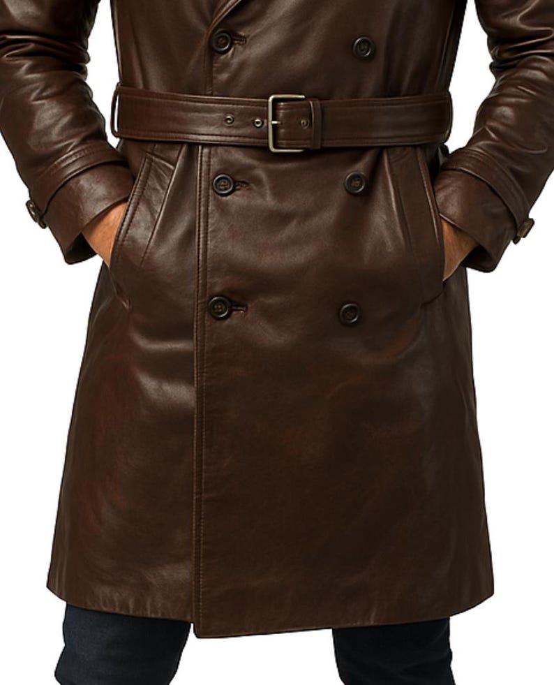 May include: A brown leather trench coat with a double-breasted design. The coat features a belt with a silver buckle, two front pockets, and button closures. The sleeves have buttoned cuffs. The person is wearing dark blue jeans.