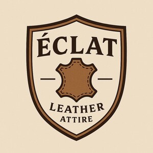 May include: A shield-shaped logo with a cream background and dark brown border. The text &Eacute;CLAT appears at the top, with LEATHER ATTIRE at the bottom. In the center, a brown leather hide icon with stitched edges is flanked by two horizontal lines.