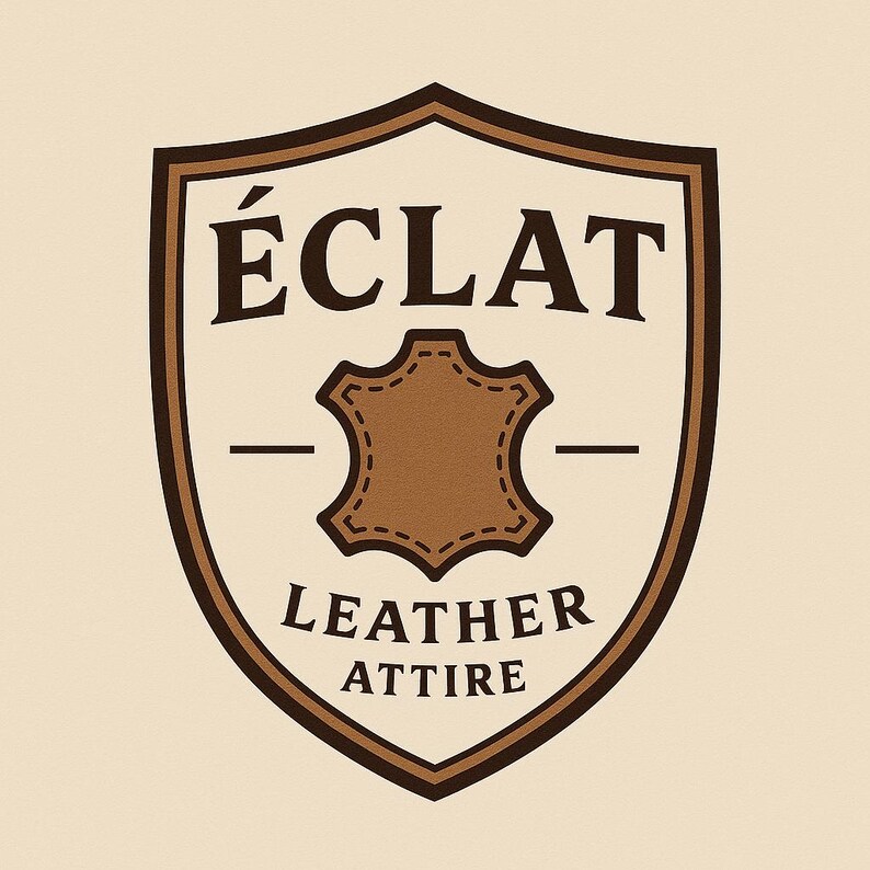 May include: A shield-shaped logo with a cream background and dark brown border. The text &Eacute;CLAT appears at the top, with LEATHER ATTIRE at the bottom. In the center, a brown leather hide icon with stitched edges is flanked by two horizontal lines.