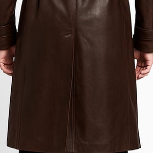 May include: A brown leather trench coat with a belt. The coat has long sleeves and a button closure. The back of the coat has a vent at the bottom. The coat is made of a smooth, shiny leather material.