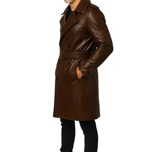 May include: A brown leather trench coat with a double-breasted design and a belt at the waist. The coat features a classic collar and button closures. The model is wearing black jeans and brown leather boots.