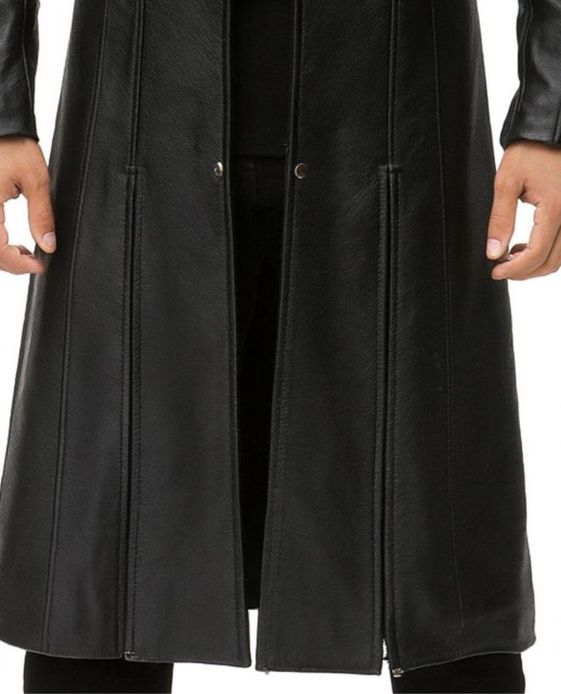 May include: A long black coat featuring a smooth texture, vertical seam detailing, and a front opening with visible metal snap buttons. The garment extends past the waist, showcasing a structured design with long sleeves and a clean, minimalist finish.
