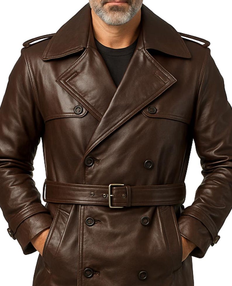 May include: A brown leather trench coat with a double-breasted design, featuring a belt with a silver buckle. The coat has a wide collar, shoulder epaulets, and multiple buttons. The person is wearing a black shirt underneath.