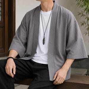 May include: A grey open-front kimono-style jacket with three-quarter length sleeves, worn over a plain white t-shirt. The outfit includes a black cord necklace featuring a dark feather pendant and a black beaded bracelet on the left wrist.