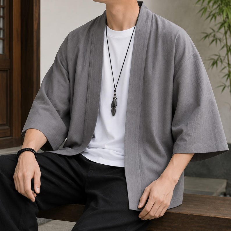 May include: A grey open-front kimono-style jacket with three-quarter length sleeves, worn over a plain white t-shirt. The outfit includes a black cord necklace featuring a dark feather pendant and a black beaded bracelet on the left wrist.