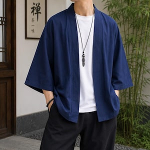 May include: A dark blue open-front kimono-style jacket with three-quarter length sleeves, worn over a plain white t-shirt and black trousers. A long black cord necklace with a metallic feather pendant hangs around the neck, and a black beaded bracelet.