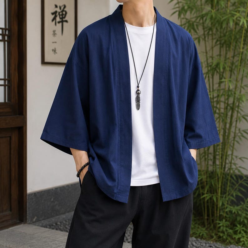 May include: A dark blue open-front kimono-style jacket with three-quarter length sleeves, worn over a plain white t-shirt and black trousers. A long black cord necklace with a metallic feather pendant hangs around the neck, and a black beaded bracelet.