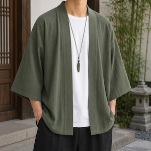 May include: An olive green open-front kimono-style cardigan with three-quarter length sleeves, worn over a plain white t-shirt and paired with black trousers. A long black cord necklace with a dark feather-shaped pendant is worn around the neck.