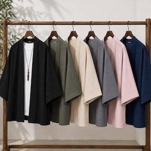 May include: A row of six open-front kimono-style jackets hanging on a wooden rack. The jackets come in black, olive green, beige, gray, light pink, and navy blue. The black jacket is layered over a white t-shirt with a long black pendant necklace.