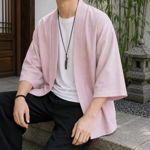 May include: A light pink open-front kimono-style jacket with three-quarter length sleeves, worn over a plain white crew-neck t-shirt. The outfit includes a black cord necklace with a feather pendant and a black beaded bracelet on the wrist.