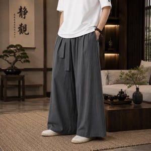 May include: A pair of dark gray wide-leg trousers featuring a high-waisted design with a matching fabric tie belt at the waist. The pants have a loose, flowing silhouette that extends to the ankles, paired with a plain white short-sleeved t-shirt.