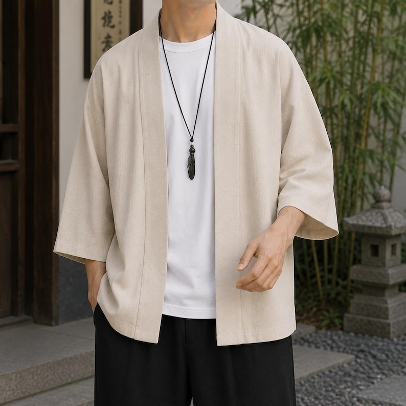 May include: A beige open-front kimono-style cardigan with three-quarter length sleeves, worn over a plain white crew-neck t-shirt. A black cord necklace with a dark feather-shaped pendant hangs around the neck, paired with solid black trousers.