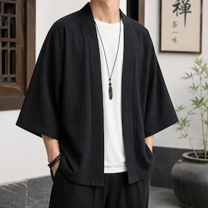 May include: A black open-front kimono-style jacket with three-quarter length sleeves, worn over a plain white t-shirt. The jacket features a simple, unfastened design with a straight hemline and a textured fabric finish, paired with a long necklace.