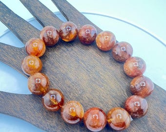 May include: A bracelet featuring a strand of polished, round beads. The beads are a mix of amber and reddish-brown hues, with a translucent quality. The bracelet is displayed on a wooden hand-shaped display.