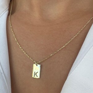 Op de afbeelding: Goudkleurige ketting met een rechthoekige hanger. De hanger heeft de letter "K" gegraveerd. De ketting wordt gedragen over een wit overhemd.
