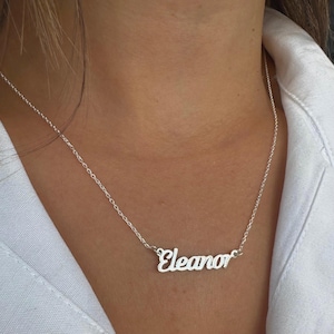 以下が含まれることがあります： 筆記体で「Eleanor」と書かれたシルバーのネックレス。白いシャツを背景に、チェーンとペンダントが見えるように首に着用されています。
