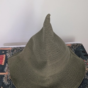 May include: Olive green witch hat with a pointed crown and wide brim. The hat is made of a knitted material. The hat is sitting on a patterned surface.