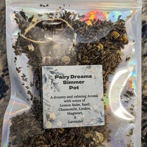 Puede incluir: Un paquete transparente e iridiscente lleno de hierbas secas. Una etiqueta dice "Fairy Dreams Simmer Pot" con una lista de ingredientes: Melisa, Albahaca, Manzanilla, Tilo, Artemisa y Lavanda. El paquete está sellado.