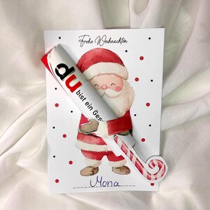 Peut inclure: Une carte de Noël avec une illustration de Père Noël tenant un sucre d'orge et un tube blanc à détails rouges. La carte porte les inscriptions "Frohe Weihnachten" et "du bist ein Geschenk".
