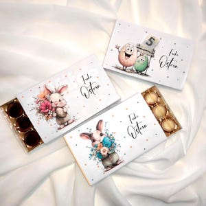Könnte beinhalten: Drei weiße Geschenkboxen mit dem Text "Frohe Ostern" und Illustrationen von Osterhasen und Eiern. Jede Box enthält Pralinen in einem goldenen Tablett. Die Boxen befinden sich auf einem weißen Stoffhintergrund.
