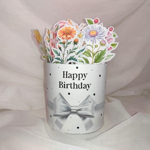 Puede incluir: Una caja de regalo cilíndrica blanca con una cinta y un lazo plateados, con el texto "Happy Birthday" y lunares negros. La caja está llena de pegatinas florales de colores, incluyendo margaritas y otras flores.