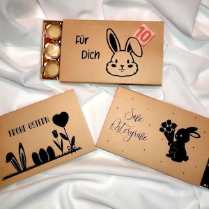 Peut inclure: Trois boîtes marron avec des motifs de Pâques. L'une présente un lapin et le texte "Für Dich". Une autre dit "Frohe Ostern" avec des oreilles de lapin et des œufs. La troisième dit "Süße Ostergrüße".