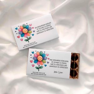 Può includere: Due scatole regalo bianche con illustrazioni di fiori colorati e testo. I fiori hanno facce sorridenti e sono circondati da cuori. Una scatola è aperta, rivelando tre oggetti rotondi marrone scuro.