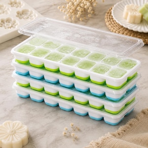 May include: A stack of four rectangular ice cube trays with clear plastic lids. Each tray features a white frame with individual green and blue silicone compartments filled with frozen green liquid, arranged in a grid pattern of four by four.