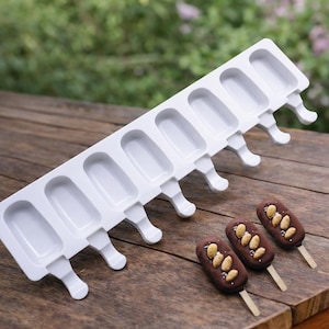 Może przedstawiać: Biała forma do lodów na patyku z ośmioma gniazdami, każde z uchwytem. Trzy oblane czekoladą lody na patyku z dodatkami migdałów znajdują się obok formy. Lody na patyku są na drewnianych patyczkach, na drewnianej powierzchni.