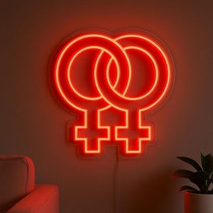 May include: A vibrant red neon sign depicting two intertwined Venus symbols, fixed to a wall. The sign is lit, producing a warm light. A sofa and a side table with a plant are visible in the background.