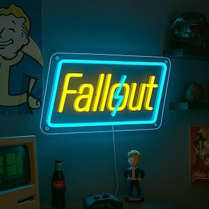 Könnte beinhalten: Ein Neonschild mit dem Wort "Fallout" in Gelb, umrandet von Blau, hängt an einer blauen Wand. Ein Vintage-Computer, ein Controller und eine Figur stehen auf einem Holztisch. Poster und Regale mit Dekoration vervollständigen den Raum.