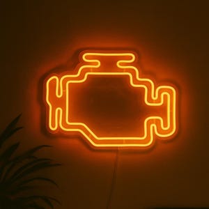 Pode incluir: Uma placa de néon laranja em forma de motor de carro. A placa está montada numa parede castanha escura. A luz de néon é brilhante e emite um brilho quente. Uma planta é visível no canto inferior esquerdo.