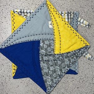 May include: A patchwork potholder with a geometric design. The potholder features sections of blue, yellow, gray, and floral fabric. The edges are stitched with black thread. A small tag is visible on the gray section.