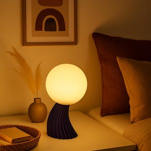 May include: A contemporary bedside lamp featuring a spherical light. The lamp has a dark purple, textured base. It sits on a white bedside table, alongside a framed abstract artwork and a small vase with dried plants.